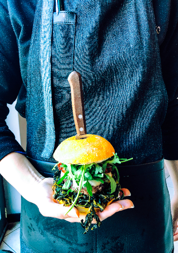 Una donna con una grembiule di jeans e pelle nera tiene sul palmo della mano un hamburger; si tratta di YODA, uno degli hamburger senza glutine di Erudito, che mostra tutti i suoi ingredienti: cicoria, hamburger, rucola, provola, ‘nduja